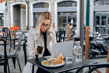 Woman working with a laptop in a cafe and eating her lunch club sandwich outdoor, freelance workplace