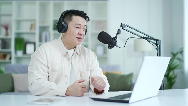 Confident asian businessman with wireless headphones recording podcast using laptop and microphone while sitting in home office. Entrepreneur communicates remotely, gives an interview via video call