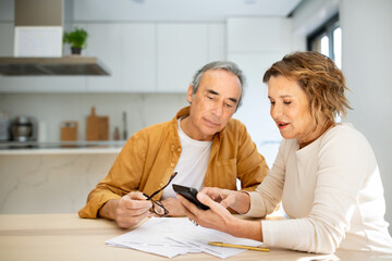 Senior couple sitting in kitchen with papers and bills and using calculator, husband and wife counting monthly expenses