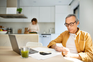 Serious aged man using cellphone, sitting at table in front of laptop in kitchen while his wife cooking on background