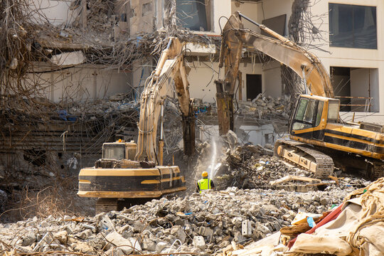 excavator equipped with a hydraulic hammer methodically demolishes an aging structure while a diligent worker sprays water to minimize dust