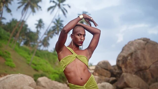 Non-binary black person in open dress, golden earrings, nose ring, bracelet, rings poses with hands to sky against palms, cliffs in tropics. Queer lgbtq fashion model wears jewelry in cocktail outfit.