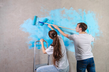 Couple in new home during repair works painting wall together. Happy family holding paint roller painting wall with blue color paint in new house. Home renovation DIY renew home concept