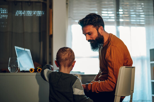 A little boy is standing next to his working father and bothering him during his father's remote job time at the home office.