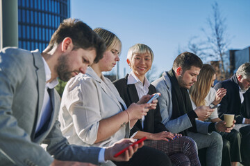 group of six businesspeople takes advantage of their break to immerse themselves in mobile technology, while one of them radiates joy as they connect with the camera