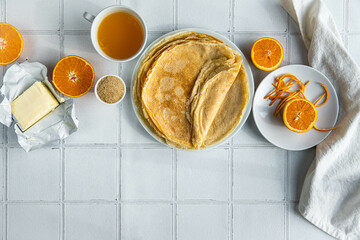 Ingredients for crepes Suzette (crepes, orange juice, brown sugar, butter) on white tiled background.