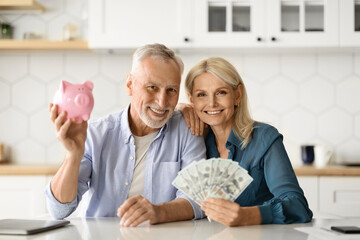 Investments Concept. Smiling Senior Couple Holding Piggybank And Dollar Cash At Home