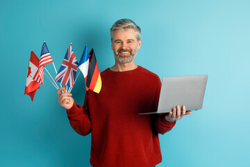 Cheerful middle aged man holding laptop and flags various countries