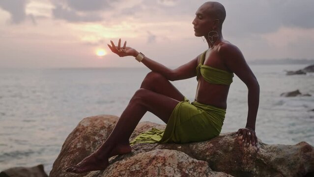 Trans sexual ethnic fashion model in long open dress, accessories in elegant posture sits on stone, looks in slow motion. Epatage gay black man in luxury gown poses above stormy ocean beach on sunset.