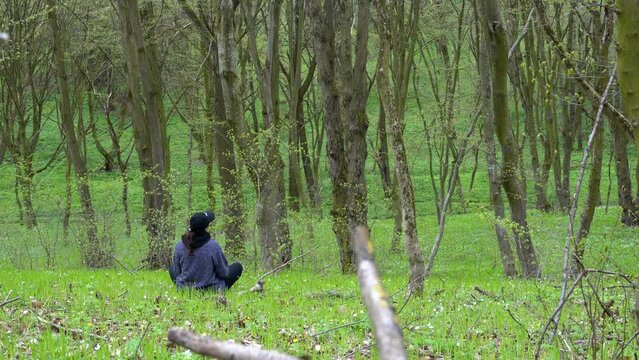 Young traveling woman sits on a green hill in the mountains and relaxing enjoying nature and birdsong. Girl meditating sitting on grass in magic forest. Healthy lifestyle, meditation concept. 4k 25FPS