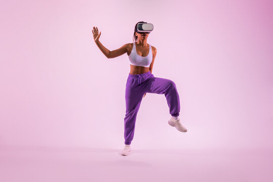 Enhancing fitness through vitual reality. Active black woman with VR glasses dancing over pink neon light background