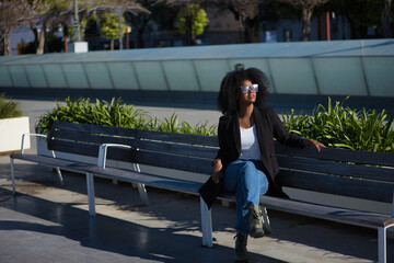 Young, beautiful, black woman with afro hair, with jacket and sunglasses sitting on a bench, receiving the sun's rays, relaxed and calm. Relaxed, calm, modern concept.