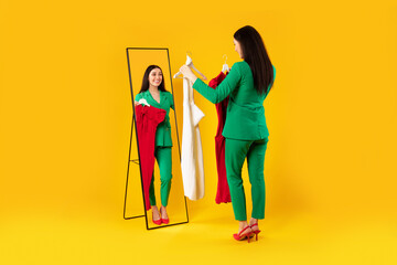 Lady shopaholic holding dresses, trying on new clothes and looking at her reflection in mirror, yellow background