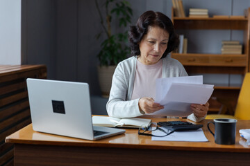 Middle aged senior woman sit with laptop and paper document, Pensive older mature lady reading paper bill pay online at home managing bank finances calculating taxes planning loan debt pension payment
