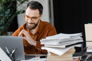 bearded businessman in stylish eyeglasses and shirt looking at laptop and smiling near smartphone with blank screen and pile of notebooks on blurred foreground in office