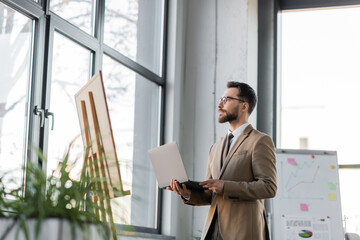 Fototapeta premium confident and fashionable bearded businessman with laptop looking away through windows near flip charts with infographics and blurred plants in contemporary office