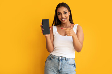 Young happy woman wearing basic clothes smiling to the camera and pointing on cellphone empty screen, posing over yellow studio background. Real people emotions