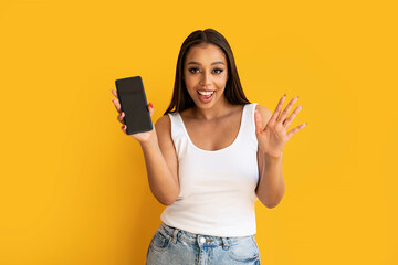 Young happy woman wearing basic clothes smiling to the camera and showing cellphone screen, posing over yellow studio background.