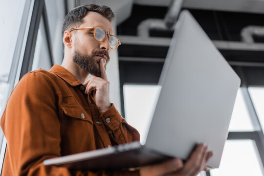 low angle view of serious bearded businessman in trendy eyeglasses and shirt looking at laptop and thinking while standing in office on blurred foreground, corporate lifestyle