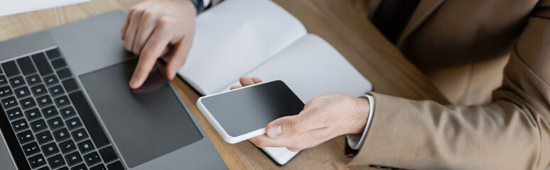 high angle view of cropped businessman in beige formal wear working on computer and holding cellphone with blank screen near empty notepad on office desk, banner