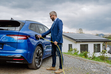 Mature businessman holding power supply cable and charging his electric car.