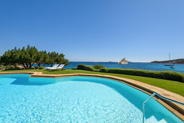 A wide-angle view of a luxury swimming pool, two lounge chairs, and a seascape with a super yacht in the distance. Italian luxury summer resort in Costa Smeralda, Sardinia