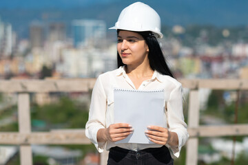 Young female architect in a hard hat on a construction site while working with a paper tablet looks...