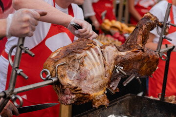 Process of cooking roasted pork on spit at summer outdoor food market, chef cutting meat - close up view. Professional cooking, cookery, gastronomy and street food concept