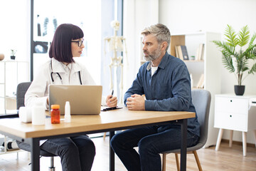 Caucasian doctor and patient sitting at table of modern medical centre and watching together at portable computer. Young specialist reading out electronic instruction for taking medication correctly.