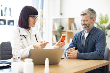 Female doctor providing expert check-up and giving medicine to middle-aged patient in hospital office. Focused man listening attentively to recommendations regarding medical history kept on laptop.