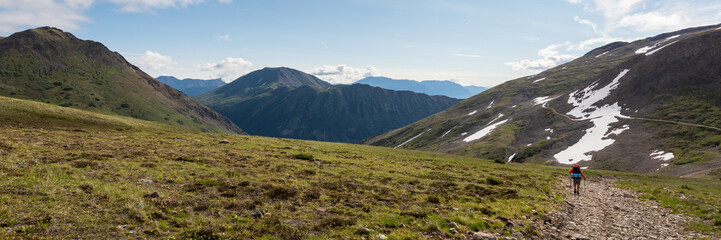Naklejka na ściany i meble One person hiking in a back country hike area along a vast, open ridge area during summer time with wilderness mountains surrounding the hiker in the wild of Alaska, Yukon, Canada. 