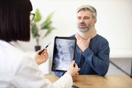 Handsome male in neck brace listening to therapist with digital x-ray images while sitting in doctor's office. Focused patient with physical sprain visiting family doctor for diagnosis in clinic.