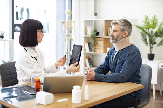 Mature bearded man in C collar talking to female physician in white coat pointing at tablet with spine scan on screen. Worried patient explaining health condition to medical person in consulting room.
