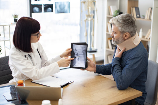Female doctor with stethoscope and male patient in neck brace sitting at writing desk with digital tablet in doctor's office. Radiologist showing cervical vertebrae causing pain on CT scan picture.