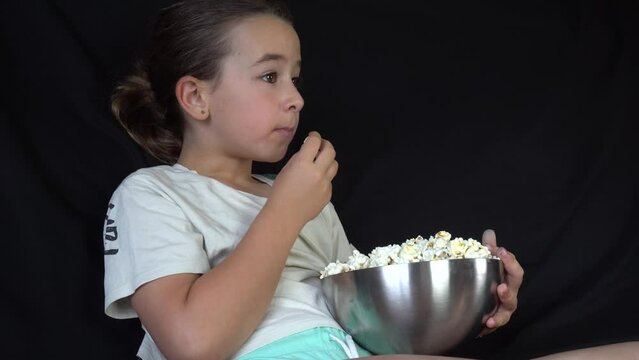 Engrossed girl sitting on sofa eating popcorn on black background while watching tv
