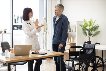 Smiling female with stethoscope clapping while happy man standing on his feet after using wheelchair. Cheerful doctor in consulting room congratulating patient with regaining mobility after accident.