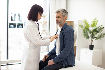Side view of focused lady in white coat using stethoscope while mature man in casual clothes...