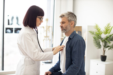 Side view of focused lady in white coat using stethoscope while mature man in casual clothes...
