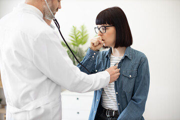 Close up view of pretty lady in eyeglasses visiting family physician for regular medical examination in clinic. Caucasian male in white coat checking lungs while patient coughing during procedure.