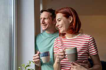 Smiling happy Caucasian couple standing near window, drinking coffee and looking outside. Positive relaxed family wife and husband enjoying weekend morning at home, planning day with partner