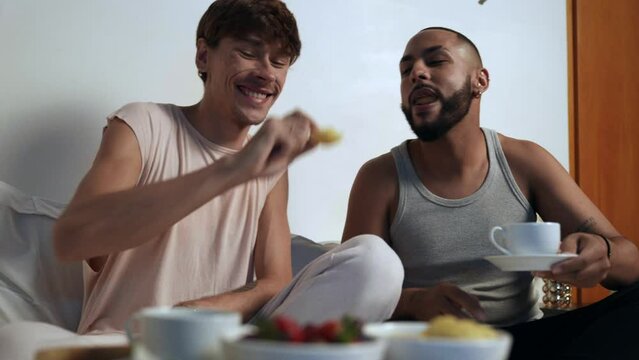 homem branco jovem brasileiro sentado na cama dando uma fruta na boca do seu namorado negro enquanto tomam caf&eacute; da manh&atilde; na cama no dia dos namorados