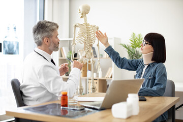 Charming woman taking seat at doctor's desk while confident man in white coat pointing at cervical...