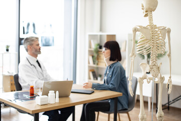 Focus on back view of teaching model located in consulting room of private practice with adults talking in background. Experts using skeleton as representation of human anatomy for patient education.