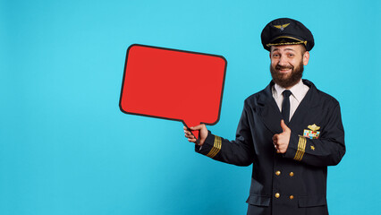 Male flying pilot holding red blank speech bubble to work on commercial flights ad in studio. Smiling positive aviator showing isolated copyspace on cardboard icon, professional captain.