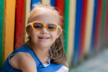 little girl with down syndrome wearing glasses for vision sits near a colored fence in a children's...