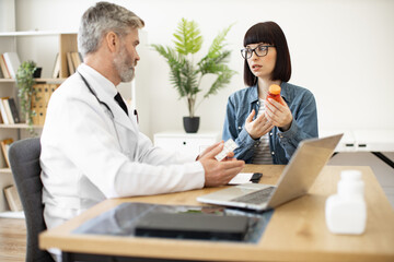 Fototapeta premium Focus on young brunette in glasses asking family physician about pills in hand while staying in consulting room. Female patient getting medical instructions about professional treatment in hospital.