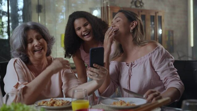 av&oacute; idosa brasileiro sentada na mesa de jantar com sua filha e neta fazendo uma v&iacute;deo chamada durante o almo&ccedil;o de dia das m&atilde;es