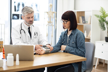 Confident bearded male with stethoscope turning on wearable tonometer applied on female wrist at doctor's office. Focused aged family physician measuring pulse rate of young caucasian woman.