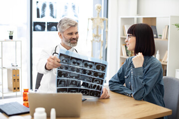 Mindful gray-haired male in lab coat holding MRI scans while brunette lady sitting next to him at desk in hospital. Family doctor examining diagnostic test results while developing treatment plan.