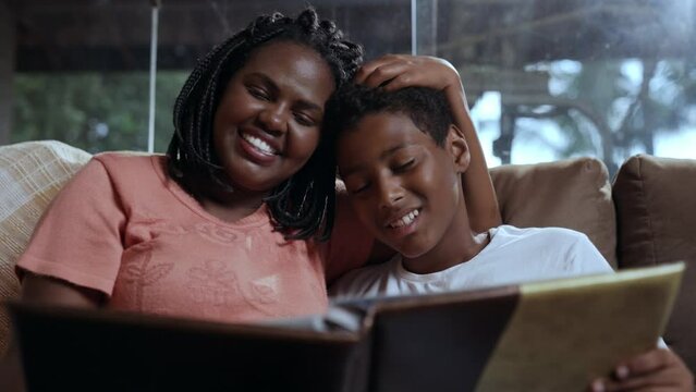 mãe brasileiro negra sorrindo enquanto mostra álbum de fotos para seu filho sentados no sofá da sala no dia das mães